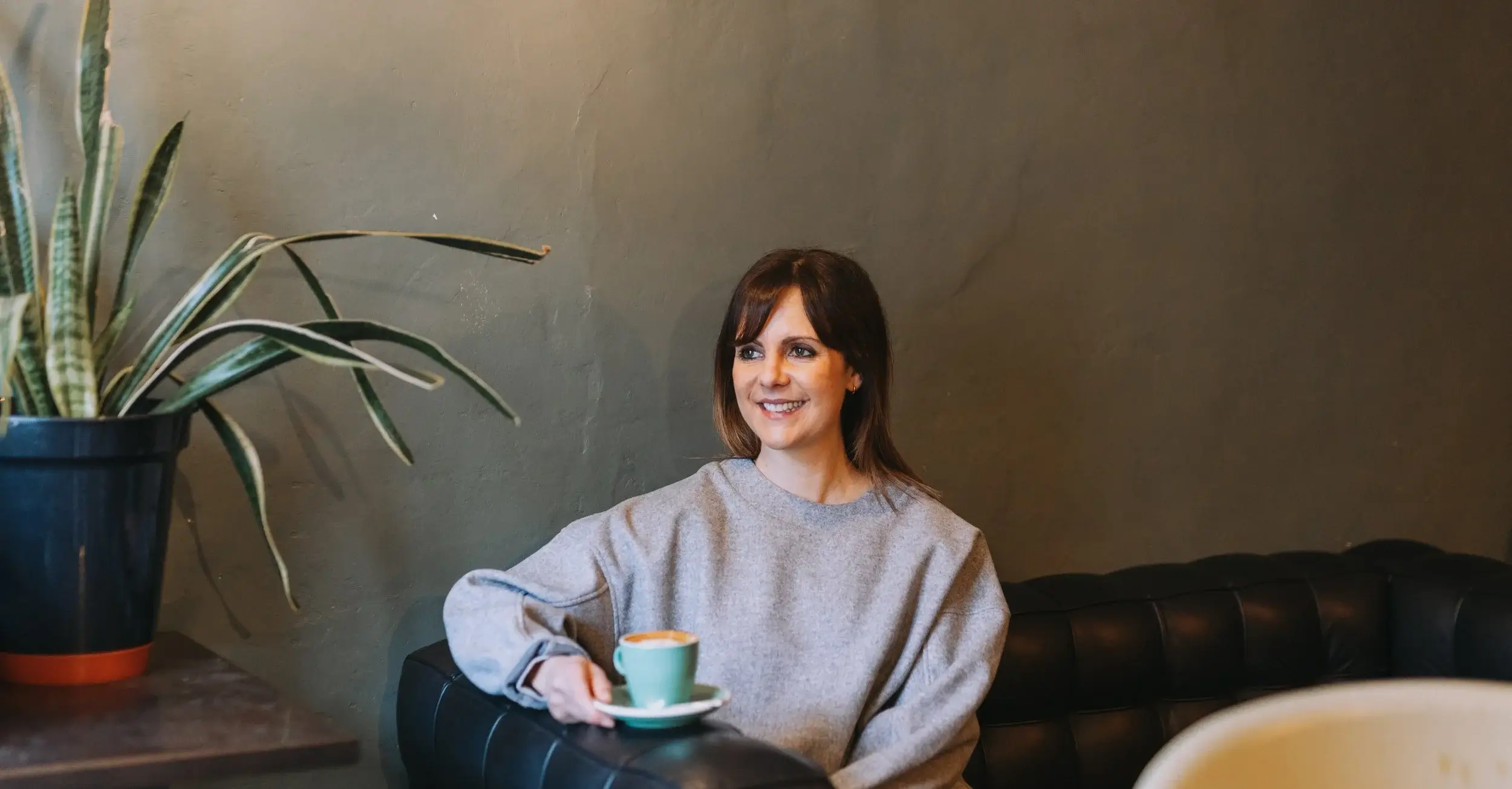 Pippa, a white woman with short dark hair, sits on a leather sofa balancing a coffee cup on the side